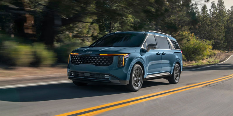 blue 2025 kia carnival driving down a wooded road